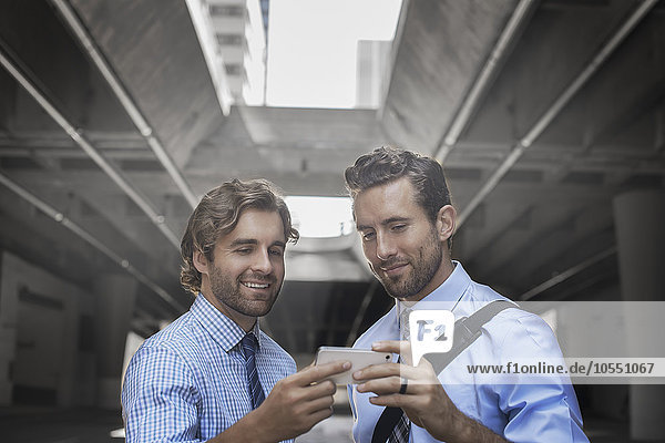 Zwei Männer in Hemd und Krawatte  die einen Selfie mit einem Smartphone nehmen  mit einem städtischen Gehweg und hohen Gebäuden im Hintergrund.