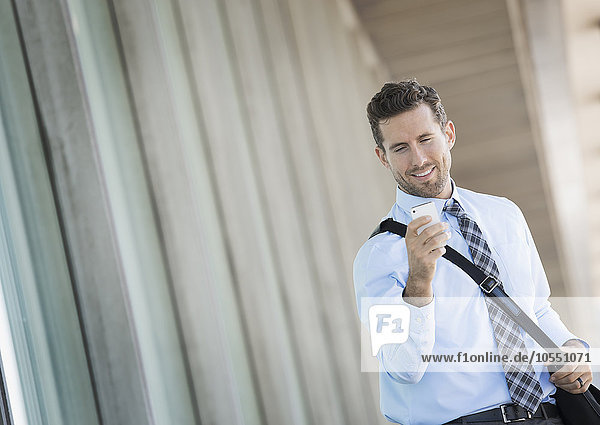 Ein Mann mit einer Umhängetasche vor einem Büro  der ein Smartphone benutzt.