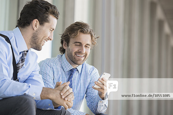 Zwei Geschäftsmänner in Hemd und Krawatte sitzen vor einem Bürogebäude und überprüfen ihre Smartphones.