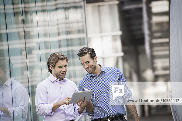 Zwei Männer stehen vor einem großen Gebäude  einer hält ein digitales Tablett in der Hand.