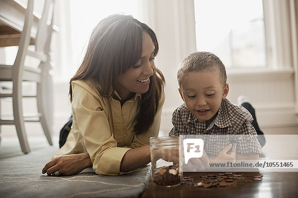 Eine Frau und ein Kind liegen auf dem Boden  spielen mit Messing- und Silbermünzen und stecken sie in ein Glasgefäß.
