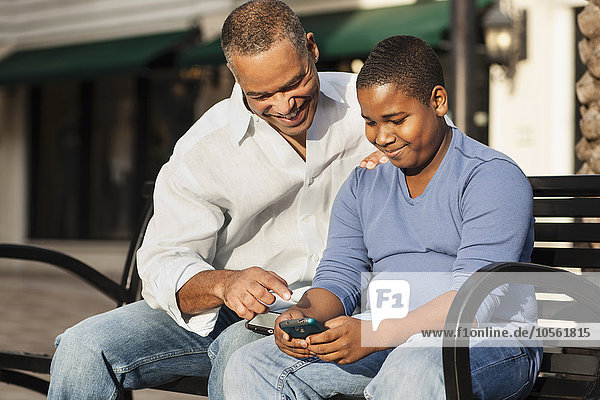 African American father and son using cell phone on bench