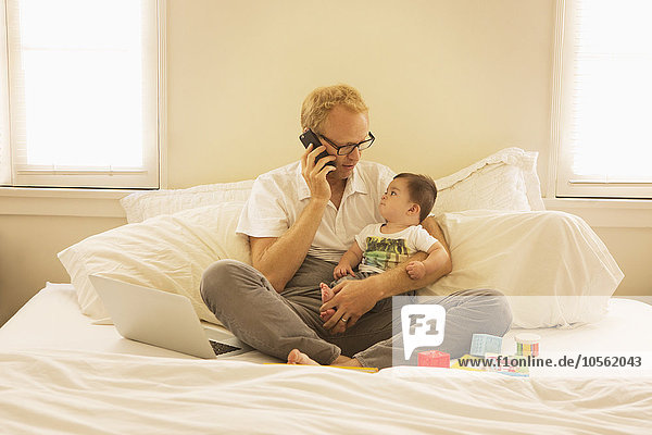 Father holding son and talking on cell phone on bed