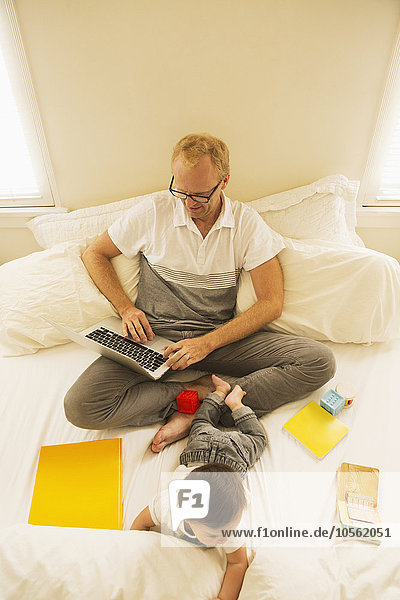 Father using laptop and playing with son on bed