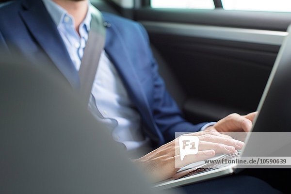 Young businessman chatting typing on laptop in back seat of car