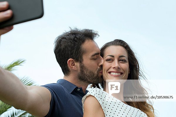 Junger Mann küsst Freundin auf der Wange für Smartphone Selfie