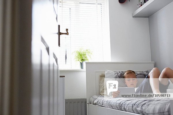 View through doorway of boy lying on bed looking down at digital tablet