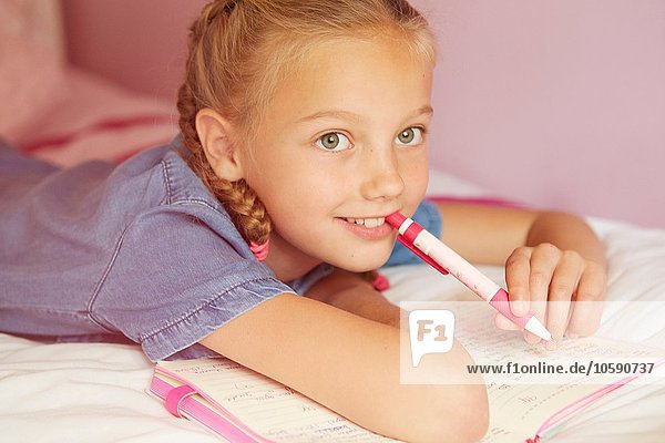 Mädchen auf dem Bett schreibt in Notizbuch  hält Stift lächelnd wegblickend