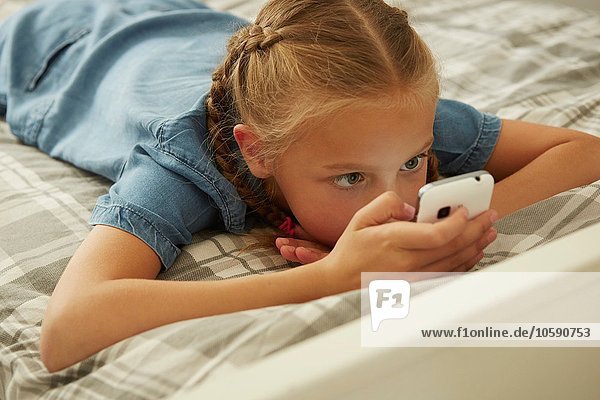 Hochwinkelansicht des auf dem Bett liegenden Mädchens mit Blick auf das Smartphone