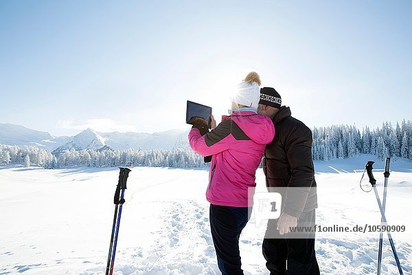 Rear view of senior couple on snowy landscape using digital tablet to take photograph of mountain range  Sattelbergalm  Tyrol  Austria