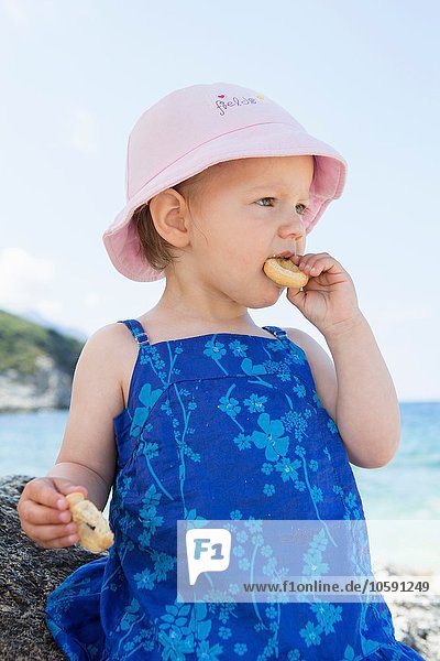 Weibliches Kleinkind mit Sonnenhut beim Donutessen am Strand