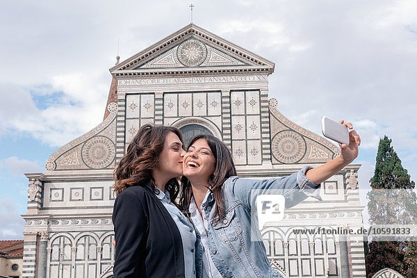 Lesbian couple using smartphone to take selfie in front of church  kiss on cheek  Piazza Santa Maria Novella  Florence  Tuscany  Italy