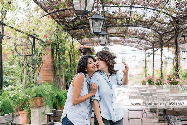 Lesbian couple in plant covered archway using smartphone to take selfie  kissing on cheek  Florence  Tuscany  Italy