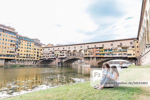 Lesbian couple sitting together on riverbank looking at digital camera in front of Ponte Vecchio and river Arno  Florence  Tuscany  Italy