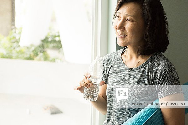 Reife Frau hält aufgerollte Yogamatte und Wasserflasche aus dem Fenster lächelnd