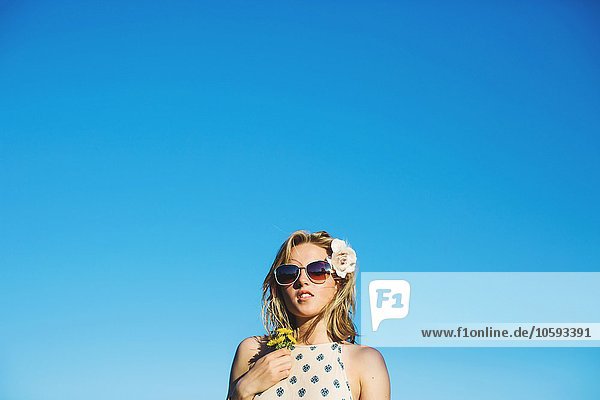 Flachwinkelansicht einer jungen Frau  die eine Sonnenbrille trägt  Blumen im Haar  die den Löwenzahn gegen den blauen Himmel halten.