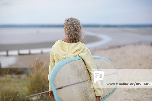 Senior woman walking towards beach  carrying surfboard  rear view