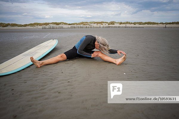 Seniorenfrau sitzt auf Sand  streckt sich  Surfbrett neben ihr