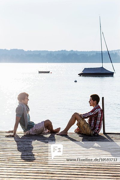 Friends sitting face to face on wood pier next to lake  Schondorf  Ammersee  Bavaria  Germany