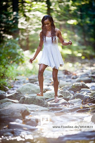 Glamorous young woman wearing white dress stepping across forest river rocks
