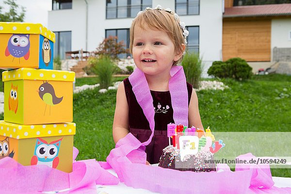 Porträt eines weiblichen Kleinkindes mit Geburtstagskuchen und rosa Bändern im Garten