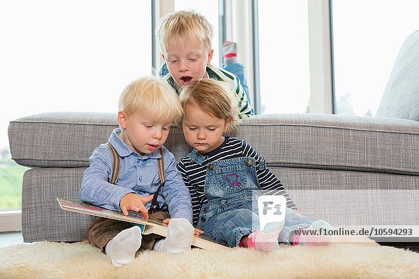 Junge und zwei Kleinkinder lesen Kinderbuch auf dem Wohnzimmerboden