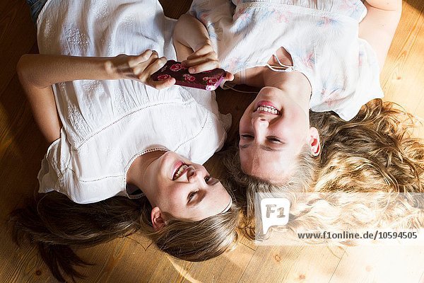 Zwei Teenager-Mädchen liegen auf dem Holzboden und lesen Smartphone-Texte.