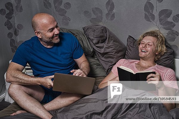 Männliches Paar sitzt im Bett und lacht beim Lesen und Benutzen des Laptops.
