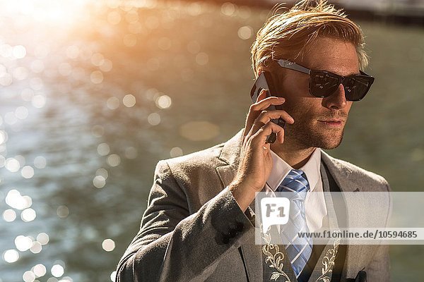 Junger Geschäftsmann mit Sonnenbrille im Gespräch auf dem Smartphone am Wasser