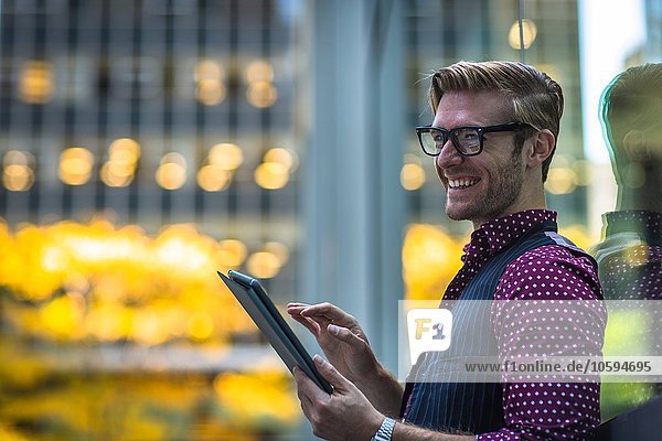 Glücklicher junger Geschäftsmann der sich mit einem digitalen Tablett gegen das Parkfenster lehnt.