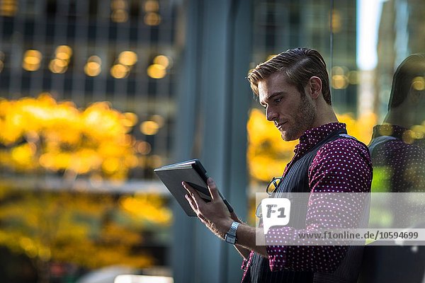 Junger Geschäftsmann der sich gegen das Parkfenster lehnt und ein digitales Tablett liest.