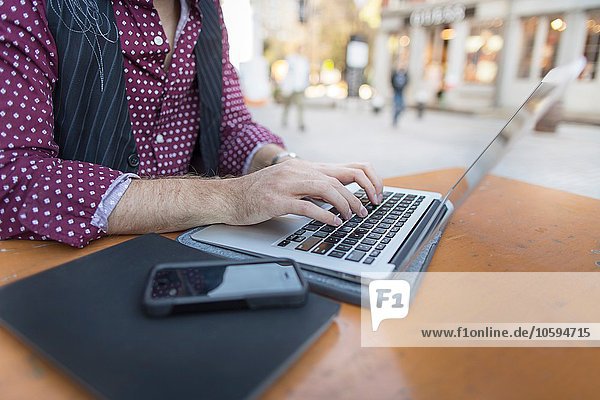 Beschnittene Ansicht des jungen Geschäftsmannes  der auf Laptop am Bürgersteig cafe schreibt