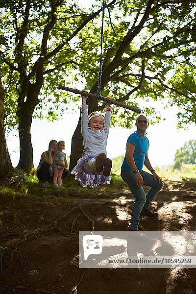 Junges Mädchen schwingt auf Baumschaukel  während ihre Familie zuschaut.