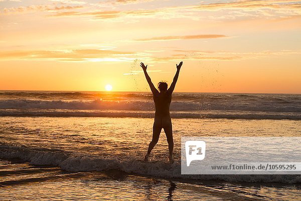 Rear view of mid adult nude womans silhouette standing in ocean at sunset  arms raised
