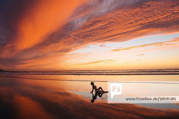 Seitenansicht der nackten mittleren erwachsenen Frauensilhouette  die am Strand sitzt und sich zurücklehnt und auf den Händen ruht und den Sonnenuntergang betrachtet.