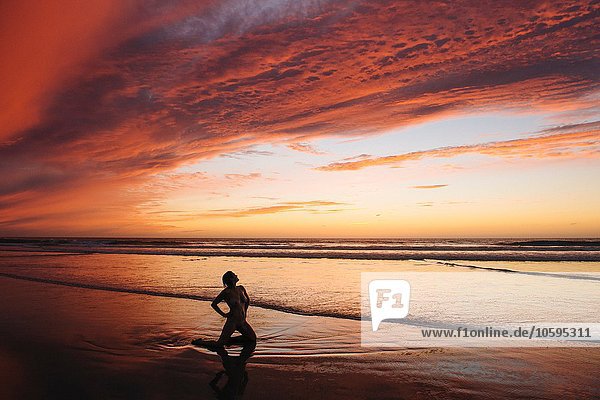 Vorderansicht der nackten mittleren erwachsenen Frauensilhouette  die bei Sonnenuntergang auf der Hüfte kniend auf dem Strand liegt.