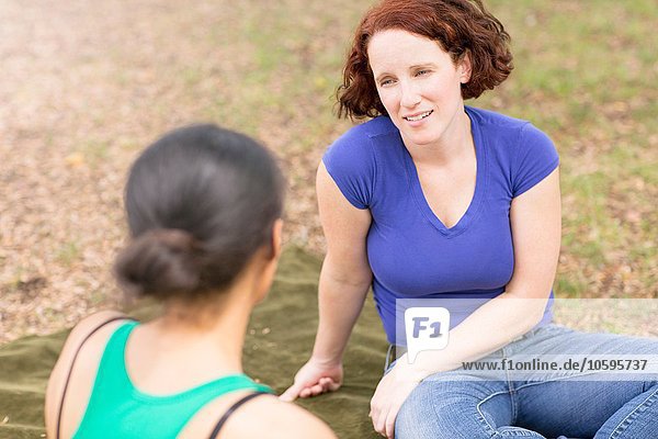 Hochwinkelansicht einer jungen Frau  die auf einer Picknickdecke sitzt und lächelnd mit einer Freundin spricht.