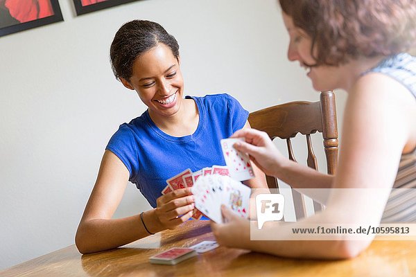 Schrägansicht von jungen Frauen  die am Esstisch sitzen und lächelnd Karten spielen.