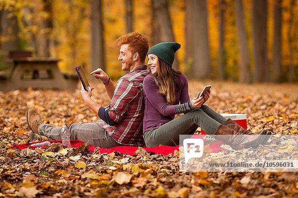 Young couple back to back using digital tablet and smartphone in forest