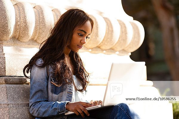 Young woman sitting leaning against stone carving looking down using laptop computer