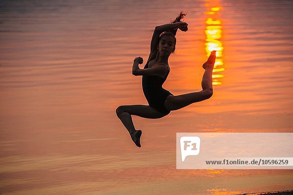 Seitenansicht des Mädchens in Silhouette am Meer bei Sonnenuntergang  Sprung in der Luft  Beine auseinander  Blick auf die Kamera