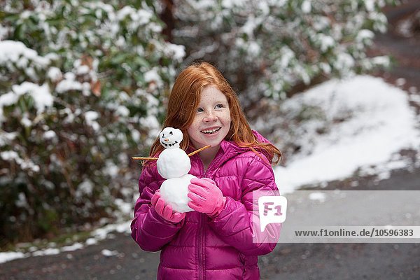 Rothaariges Mädchen vor schneebedeckten Bäumen  mit rosa Handschuhen und gepolsterter Jacke  die einen Schneemann hält  der lächelnd wegsieht.