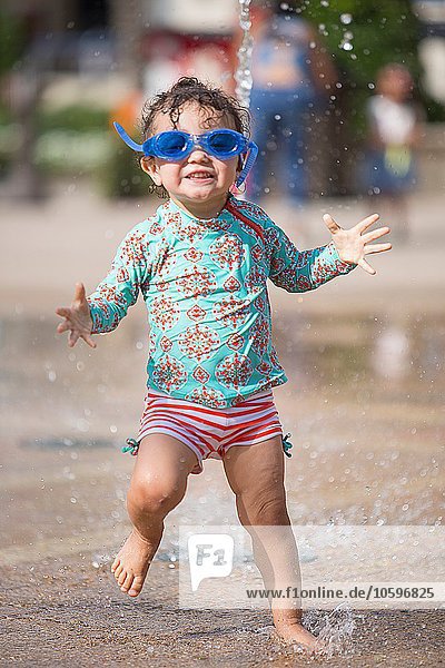 Girl wearing swimming goggles playing in water fountain  looking at camera smiling