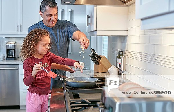 Father helping daughter cook on hob in kitchen  sprinkling cheese