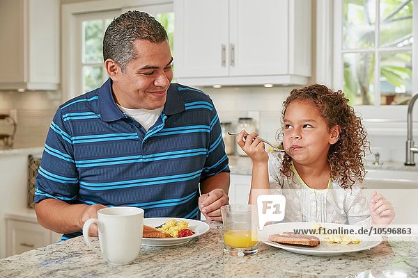 Father and daughter side by side at kitchen counter eating breakfast smiling
