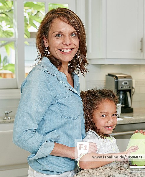 Seitenansicht der Mutter hinter der Tochter in der Küche mit lächelndem Blick auf die Kamera