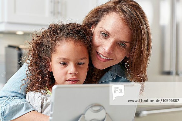 Mother with arm around daughter using digital tablet looking down smiling