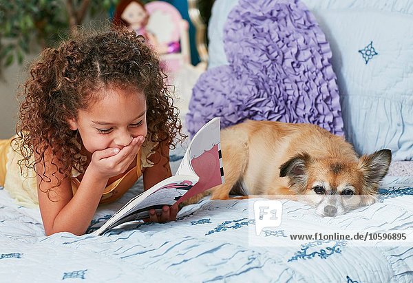 Girl and dog lying on bed reading book  hand on mouth laughing