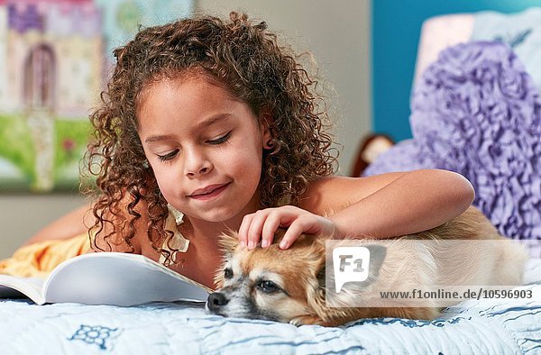 Girl lying on bed reading book stroking dog