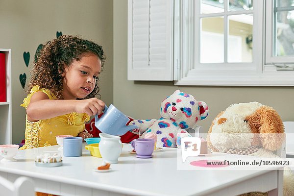 Mädchen im Spielzimmer sitzt am Tisch und serviert Tee vom Spielzeugtee bis zum Plüschtier.